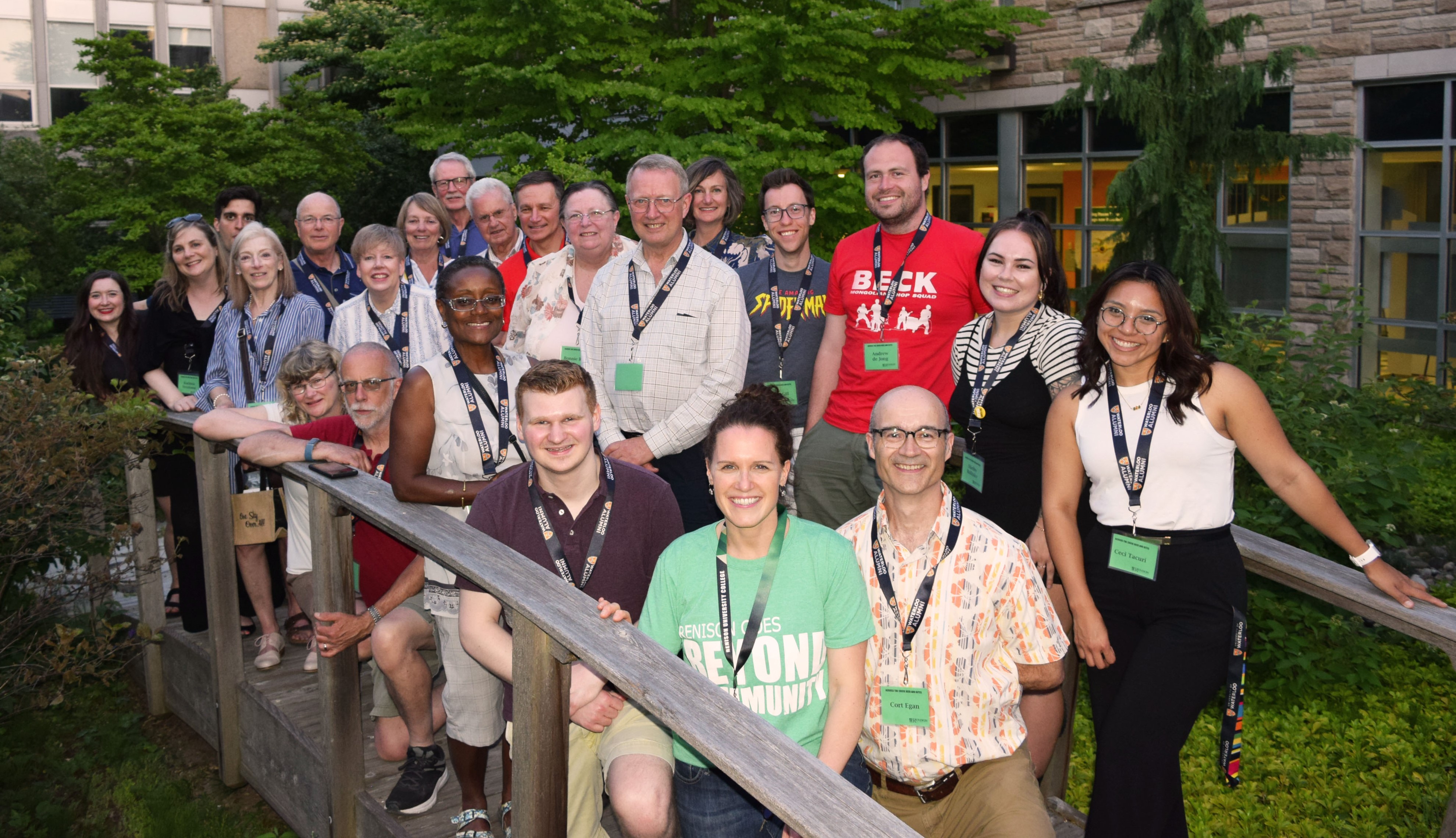 Group of alumni stand on the bridge in the Renison courtyard. Everyone is smiling and all are wearing name tags from the event.